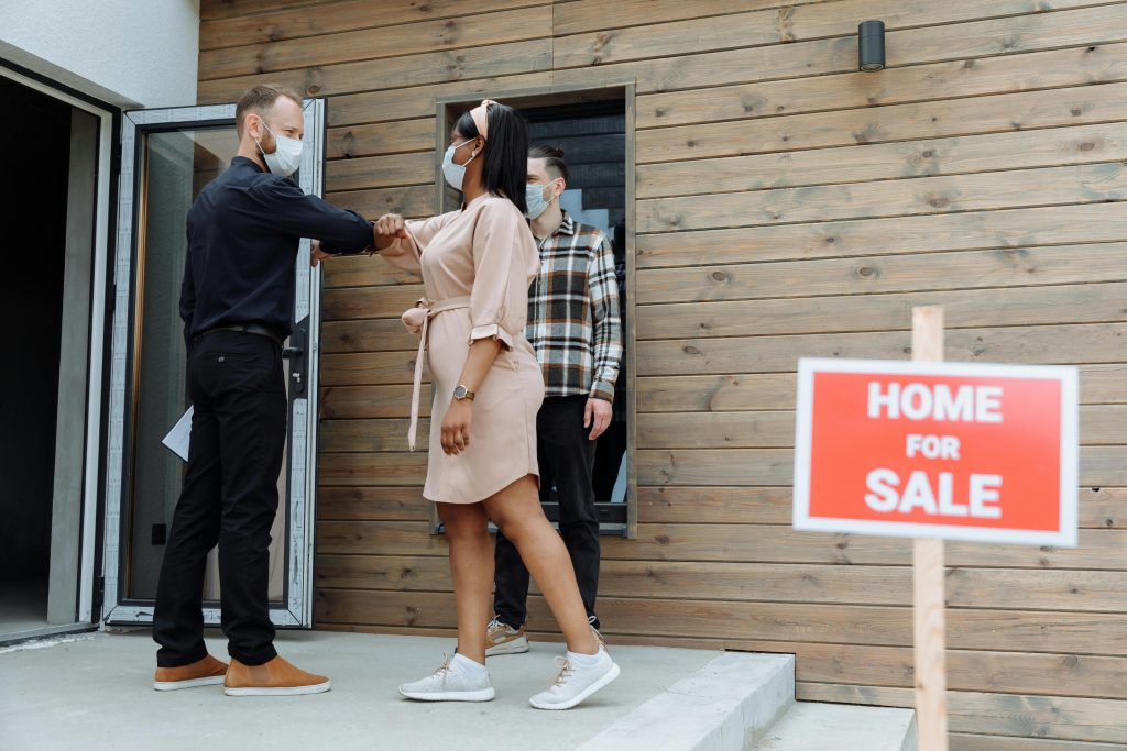 Masked real estate agent greets clients with elbow bump outside a home for sale.